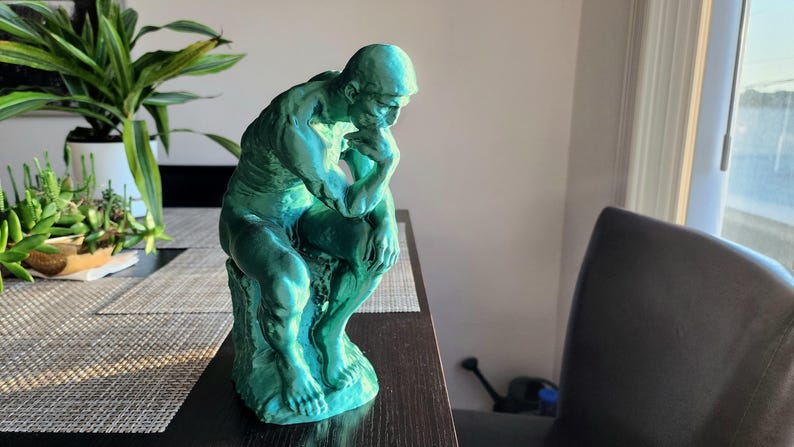 May include: A teal-colored sculpture of a seated figure, reminiscent of Rodin's "The Thinker." The statue is positioned on a dark surface, with a plant and a window in the background. The sculpture is approximately 10 inches tall.