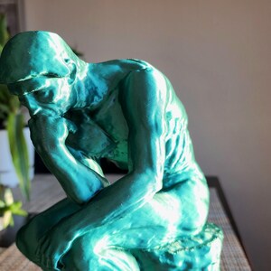 May include: A teal-colored sculpture of a seated figure, with the head resting on a hand, in a contemplative pose. The statue is on a table, with plants in the background. The sculpture is a representation of The Thinker.
