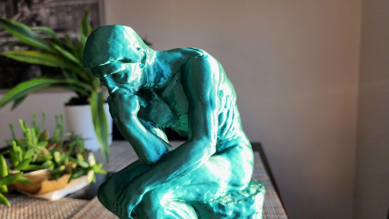 May include: A teal-colored sculpture of a seated figure, with the head resting on a hand, in a contemplative pose. The statue is on a table, with plants in the background. The sculpture is a representation of The Thinker.