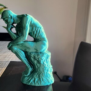 May include: A teal-colored sculpture of Rodin's The Thinker. The figure is seated, with a hand resting on the chin, and is atop a textured base. The sculpture is placed on a dark surface near a window.