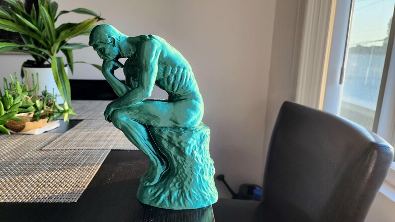 May include: A teal-colored sculpture of Rodin's The Thinker. The figure is seated, with a hand resting on the chin, and is atop a textured base. The sculpture is placed on a dark surface near a window.