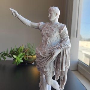 May include: A white, textured statue of a Roman figure in military armor, standing on a rectangular base. The figure has one arm extended outward, pointing, and is draped in a long, flowing garment that falls to the knees, revealing bare legs.