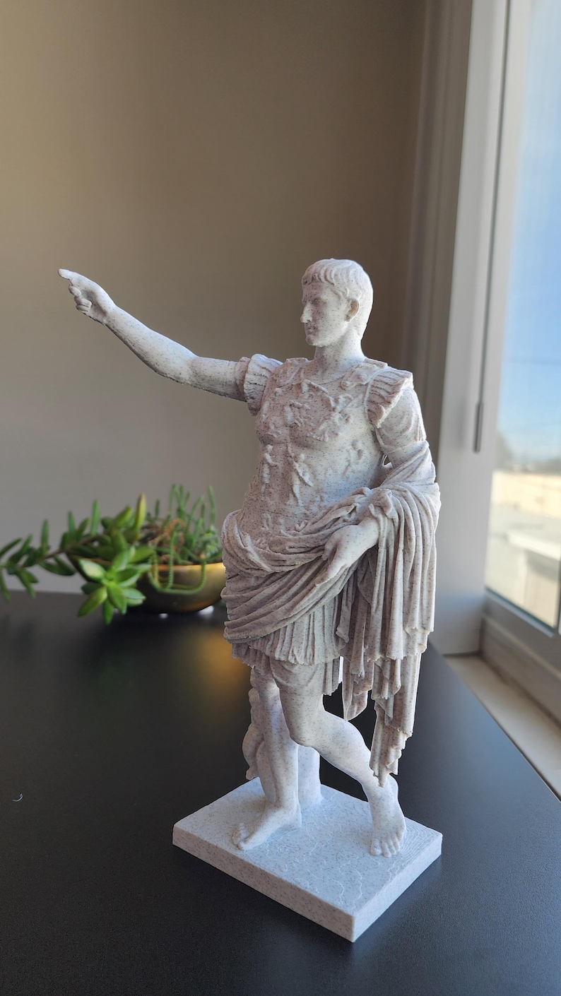 May include: A white, textured statue of a Roman figure in military armor, standing on a rectangular base. The figure has one arm extended outward, pointing, and is draped in a long, flowing garment that falls to the knees, revealing bare legs.