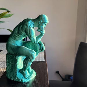 May include: A teal-colored sculpture of a seated man in a thoughtful pose. The figure is resting his chin on his hand, with his elbow on his knee. The sculpture is on a textured base, and the figure is in a room with plants.