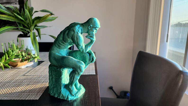 May include: A teal-colored sculpture of a seated man in a thoughtful pose. The figure is resting his chin on his hand, with his elbow on his knee. The sculpture is on a textured base, and the figure is in a room with plants.