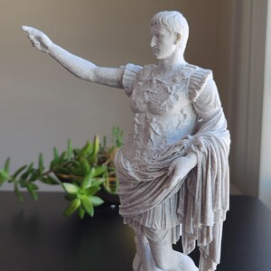May include: A white, textured statue of a Roman figure standing on a rectangular base. The figure wears detailed armor and a draped garment, with one arm extended outward and pointing. The sculpture features a matte finish and intricate carvings.