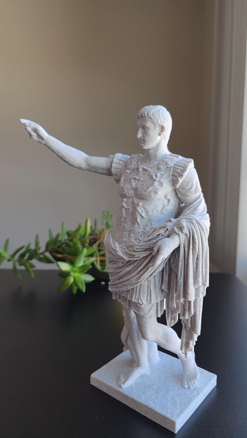 May include: A white, textured statue of a Roman figure standing on a rectangular base. The figure wears detailed armor and a draped garment, with one arm extended outward and pointing. The sculpture features a matte finish and intricate carvings.