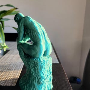 May include: A teal-colored sculpture of a seated figure, reminiscent of Rodin's "The Thinker." The statue is positioned on a textured base, with the figure's back and form clearly visible. The sculpture is placed on a dark table near a window.