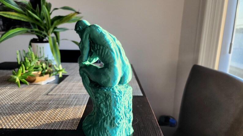 May include: A teal-colored sculpture of a seated figure, reminiscent of Rodin's "The Thinker." The statue is positioned on a textured base, with the figure's back and form clearly visible. The sculpture is placed on a dark table near a window.