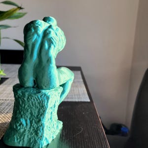 May include: A teal-colored sculpture of a seated figure, possibly a representation of Rodin's "The Thinker." The sculpture is mounted on a textured base, and the figure is posed with its back to the viewer. The sculpture is on a dark table.