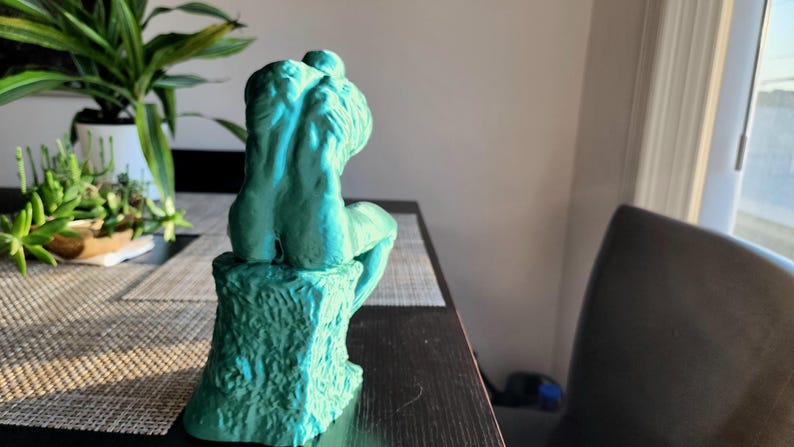 May include: A teal-colored sculpture of a seated figure, possibly a representation of Rodin's "The Thinker." The sculpture is mounted on a textured base, and the figure is posed with its back to the viewer. The sculpture is on a dark table.