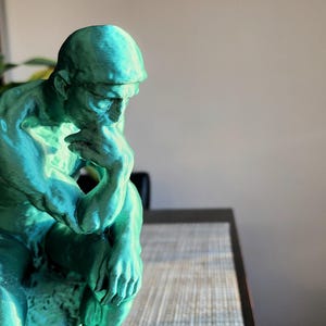 May include: A teal-colored sculpture of a man in a seated position, with his chin resting on his hand, in a contemplative pose. The statue has a textured surface and is placed on a table. The background includes plants and a framed picture.