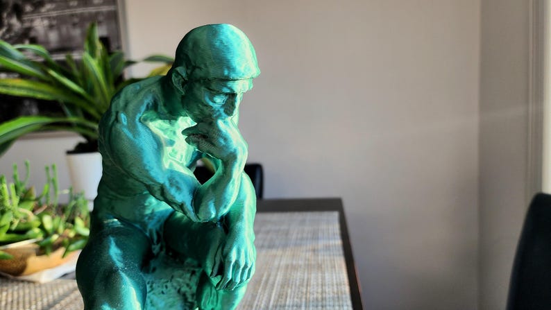 May include: A teal-colored sculpture of a man in a seated position, with his chin resting on his hand, in a contemplative pose. The statue has a textured surface and is placed on a table. The background includes plants and a framed picture.