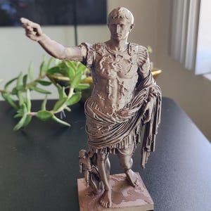 May include: A bronze-colored figurine of Augustus of Prima Porta standing on a square base. The figure wears detailed Roman military armor and a draped toga, with the right arm extended forward and a small figure positioned near the right leg.