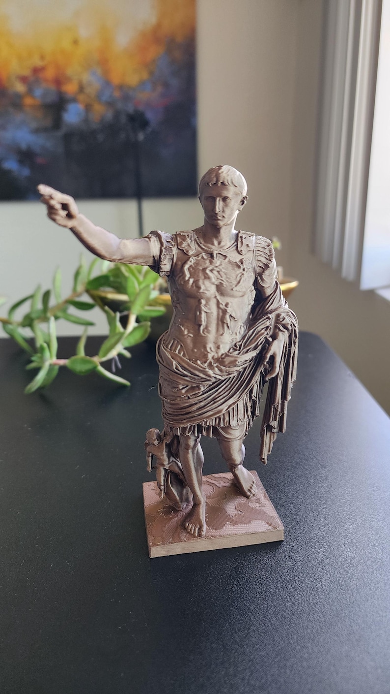 May include: A bronze-colored figurine of Augustus of Prima Porta standing on a square base. The figure wears detailed Roman military armor and a draped toga, with the right arm extended forward and a small figure positioned near the right leg.
