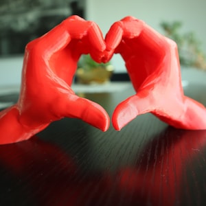 May include: Two red hands forming a heart shape. The hands are made of a smooth, solid material and are positioned on a black surface.