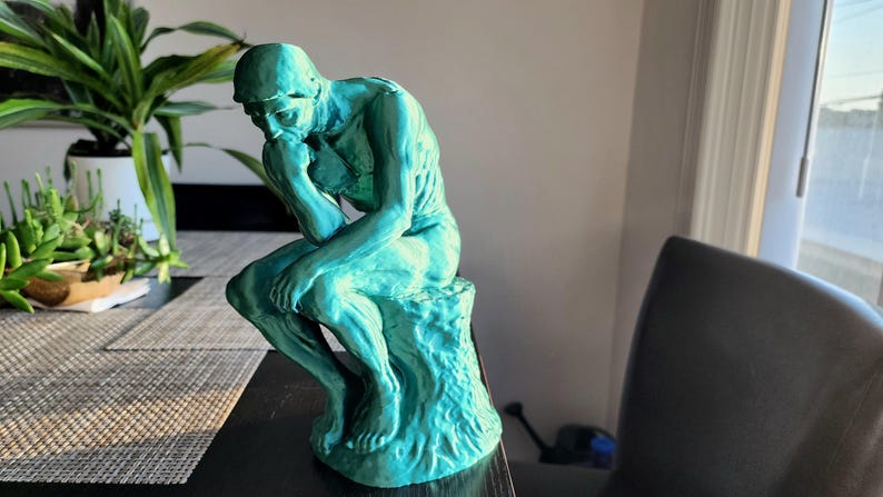 May include: A teal-colored sculpture of a seated figure in a contemplative pose, resting his chin on his hand. The statue is on a textured base, with a naturalistic, rough appearance. The sculpture is indoors, with plants in the background.