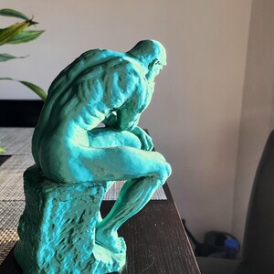May include: A teal-colored sculpture of a seated figure, reminiscent of Rodin's "The Thinker." The statue is positioned on a dark surface, with a detailed depiction of the human form. The sculpture is a decorative item.