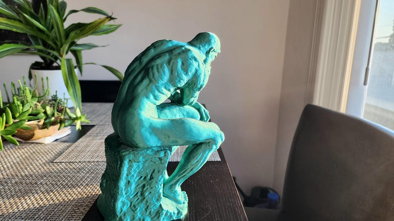 May include: A teal-colored sculpture of a seated figure, reminiscent of Rodin's "The Thinker." The statue is positioned on a dark surface, with a detailed depiction of the human form. The sculpture is a decorative item.