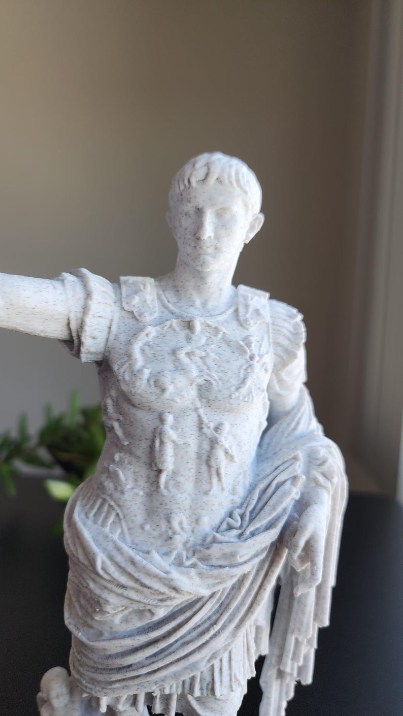 May include: A white and grey speckled statue of a Roman figure wearing detailed armor with embossed imagery on the chest plate. The figure has short hair and is draped in a textured garment that falls over the shoulder and across the torso.