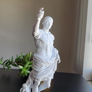 May include: A white, textured statue of a Roman figure in military armor with a draped garment around the waist. The figure stands on a square base with one arm raised and a small winged cherub figure positioned near the lower leg area.