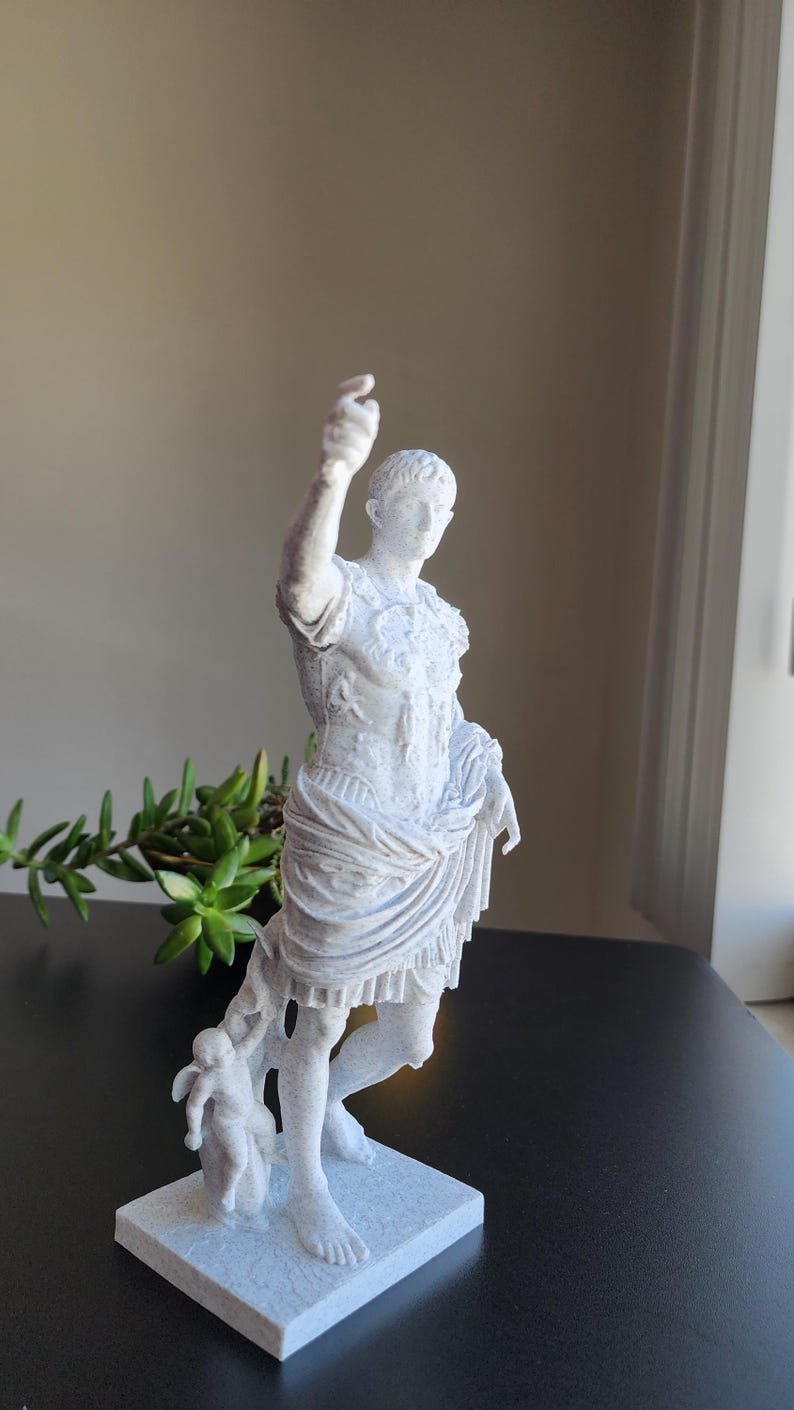 May include: A white, textured statue of a Roman figure in military armor with a draped garment around the waist. The figure stands on a square base with one arm raised and a small winged cherub figure positioned near the lower leg area.