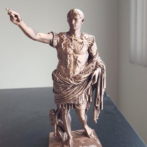 May include: A bronze-colored statue of a Roman figure standing on a square base. The figure wears detailed armor with embossed imagery, a draped tunic, and has one arm extended outward. A small cherub-like figure stands near the right leg.