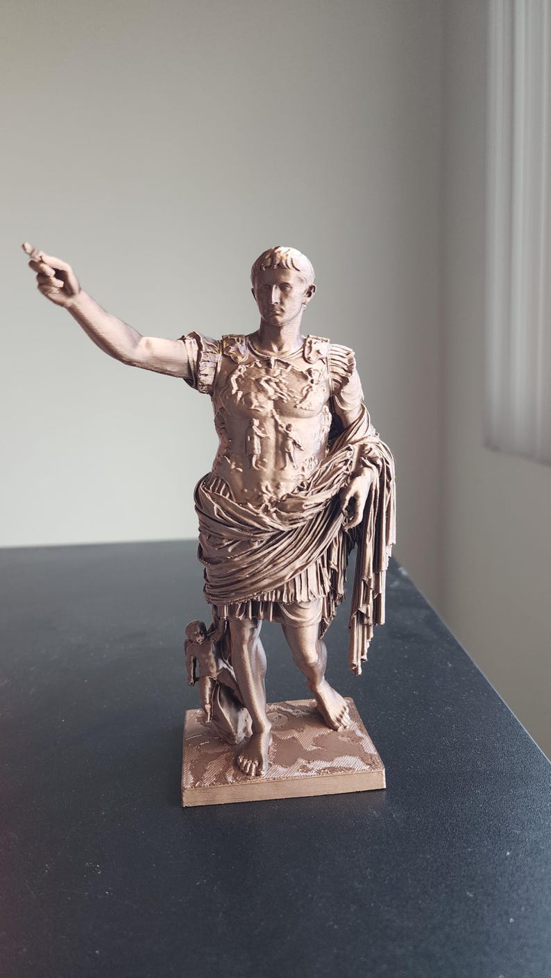 May include: A bronze-colored statue of a Roman figure standing on a square base. The figure wears detailed armor with embossed imagery, a draped tunic, and has one arm extended outward. A small cherub-like figure stands near the right leg.