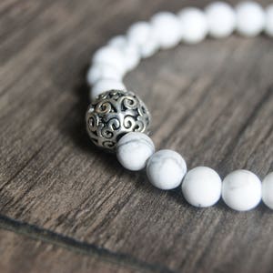 May include: White beaded bracelet with a silver filigree accent bead. The bracelet features round, white beads with a marbled texture. The silver bead has an ornate, openwork design. The bracelet is displayed on a wooden surface.
