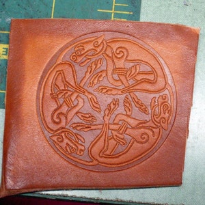 May include: A brown leather piece with a circular design of four stylized animals intertwined. The design is embossed into the leather.