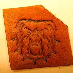 May include: A brown leather patch with a detailed embossed image of a bulldog's head with a spiked collar.