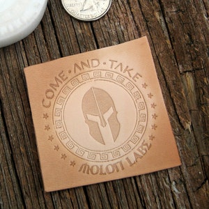 May include: A brown leather patch with the text "COME AND TAKE" and a circle design with a Spartan helmet inside. The text "MOLOTI LAB" is below the circle design.