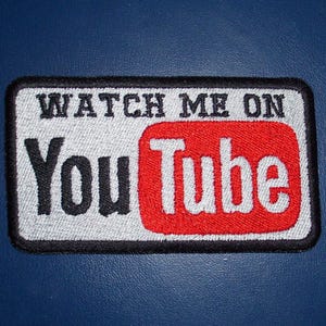 May include: A black and white embroidered patch with the text "Watch Me On YouTube" in white lettering on a gray background. The YouTube logo is red with white lettering.