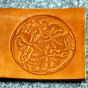 May include: A brown leather square with a circular design of four intertwined animals. The design is embossed into the leather and has a Celtic knot-like pattern.