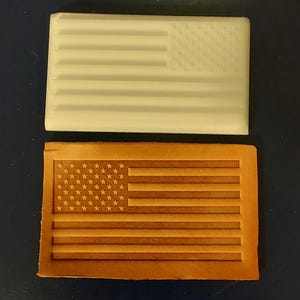 May include: Two rectangular objects, one cream-colored and the other brown, each feature an embossed American flag design. The cream-colored object is smooth, while the brown object appears to be made of leather. The flag design includes stars and stripes.