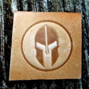 May include: A brown leather patch with a white Spartan helmet design inside a circle.