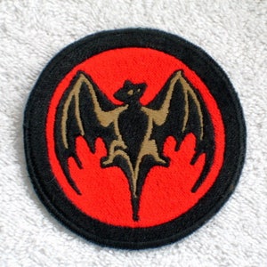 May include: A black and red embroidered patch with a brown bat in the center. The bat is inside a red circle with a black outline. The circle is inside a black circle with a white outline.