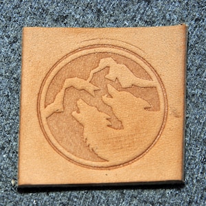 May include: A brown leather patch with a circular design featuring three wolf silhouettes howling at the moon. The wolves are facing the viewer and are surrounded by a mountain range.