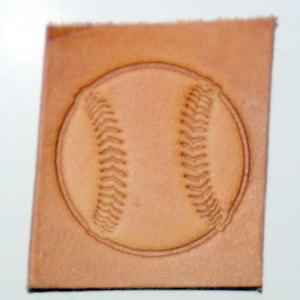 May include: A close-up of a leather stamp with a baseball design. The stamp is light brown and has a raised design of a baseball with stitching.