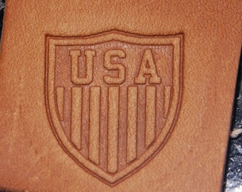 May include: A brown leather patch with a shield-shaped design. The shield has the word "USA" in white lettering and vertical stripes.