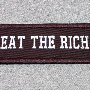 May include: A brown fabric patch with white embroidered text that reads "EAT THE RICH".