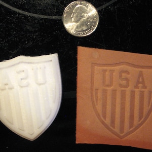May include: Two close-up images of a shield-shaped stamp. The first image shows a white plastic stamp with the letters "A2U" in the center. The second image shows a brown leather stamp with the word "USA" in the center.
