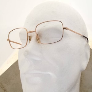 Puede incluir: Gafas rectangulares doradas con lentes transparentes. Las monturas tienen un diseño fino y delicado. Las gafas se muestran en una cabeza de maniquí blanca, mostrando la forma y el estilo de las gafas. Las patillas tienen una punta marrón oscuro.