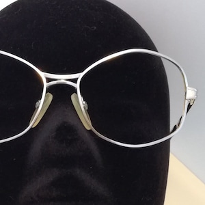 Puede incluir: Gafas con montura plateada y lentes ovaladas. Las gafas tienen una montura metálica fina y un puente con almohadillas nasales. Las lentes están tintadas y las gafas se exhiben en una cabeza de maniquí negra.