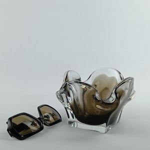 May include: A pair of square-framed sunglasses with brown lenses and a dark frame, next to a decorative glass bowl. The bowl has a unique, organic shape with a smoky brown hue, and a clear center. The items are set against a white background.