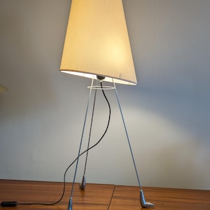 May include: A floor lamp featuring a tall, off-white, tapered fabric shade supported by three thin, silver-toned metal legs. Each leg terminates in a small, foot-shaped base. A black electrical cord hangs from the center, and the lamp is illuminated.