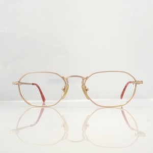 May include: Gold-toned eyeglasses with a thin metal frame and oval lenses. The temples have red accents. The glasses are reflected on a white surface.