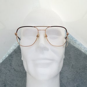 Könnte beinhalten: Goldfarbene Metallbrille mit braunen Kunststoffakzenten an den Bügeln. Die Brille hat eine rechteckige Form und klare Gläser.
