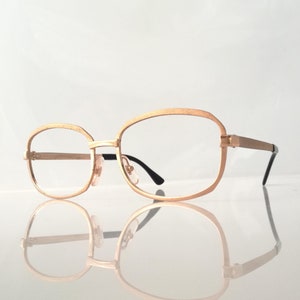 May include: Gold-tone metal eyeglasses with a square frame and clear lenses. The glasses are reflected in a white surface.
