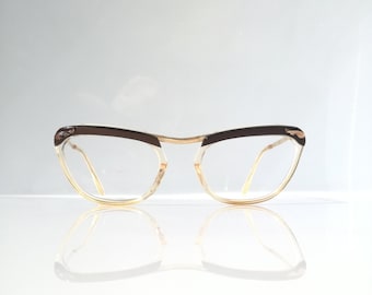 May include: A pair of vintage eyeglasses with a cat-eye shape. The frames feature a brown upper rim and temples, with a gold-colored bridge and arms. The lenses are clear, and the glasses are reflected on a white surface.
